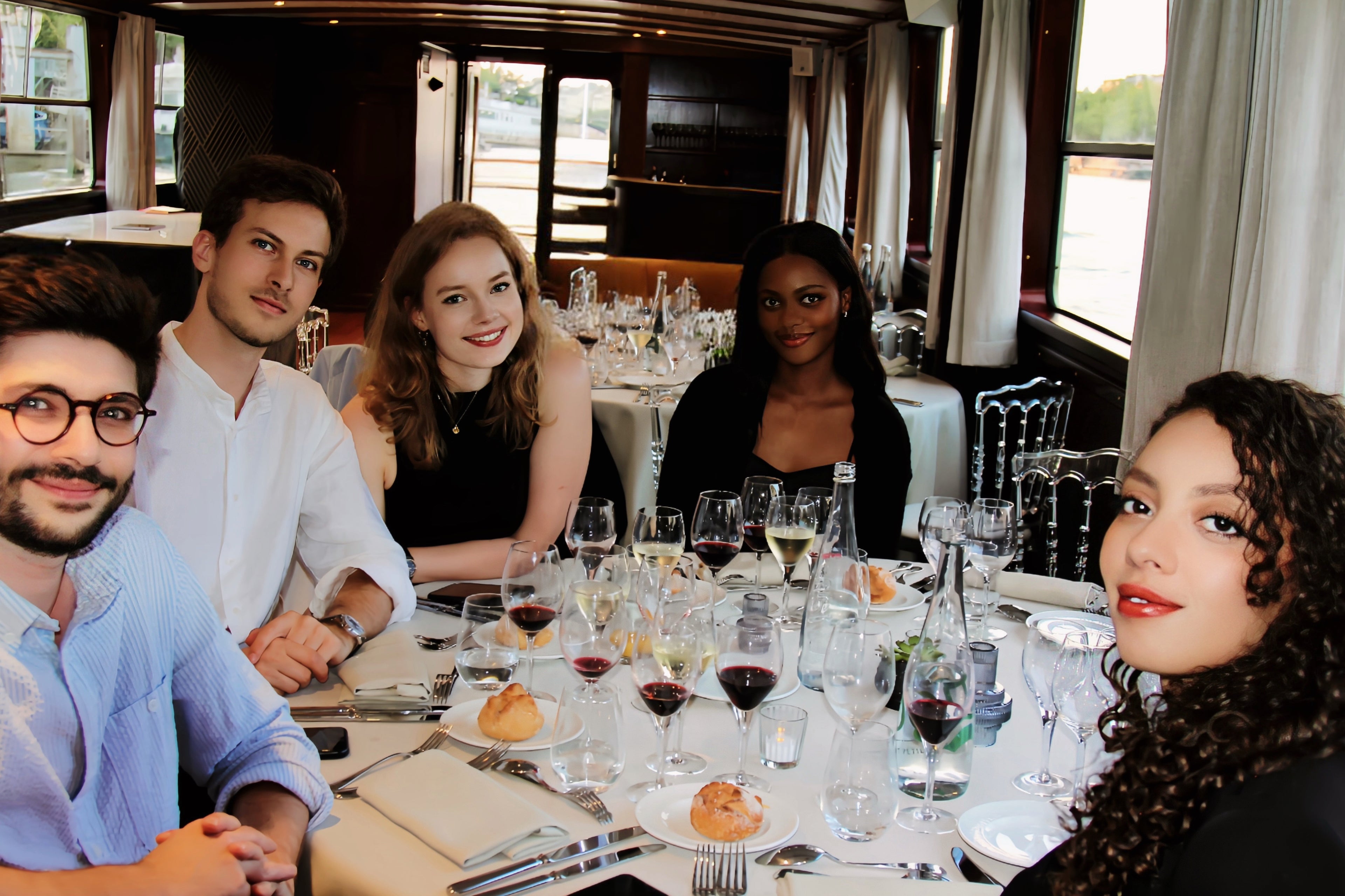Group of people sitting around a table and smiling with wine glasses and food, on a luxury yacht in Paris.